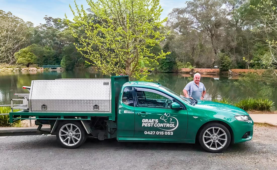 Grae, the owner of Grae’s Pest Control, stands beside his branded green utility vehicle in Mittagong. The vehicle displays the company name and phone number and is parked near a body of water with trees in the background.